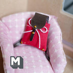 1:12 scale miniature drawstring backpack in red with a brown flap and white cord, shown on a pink armchair