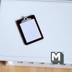 1:12 scale miniature clipboard in black with a silver clip and blank white note pad, shown on a white table