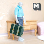 1:12 scale miniature faux leather suitcase in green with light straps, non opening, shown carried by an artist figure