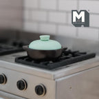 1:12 scale miniature clay pot with lid in black with a mint lid and side handles, shown on a silver stove