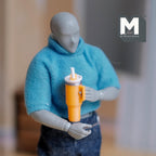 1:12 scale miniature tumbler with straw and handle in orange, held by a posable figure in a blue sweater