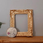 1:12 scale miniature ornate rectangular picture frame in gold with an open center, shown on a shelf beside a coin