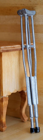 1:12 scale miniature metal underarm crutches in silver with gray pads, set of 2, shown leaning against a wood dresser
