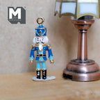 1:12 scale miniature metal nutcracker soldier ornament in blue with gold trim and a hanging loop, shown beside a table lamp