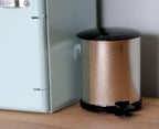 1:12 scale miniature pedal trash can with lid in rose gold and black, shown on a wood floor beside a mint green fridge