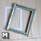 1:12 scale miniature ornate picture frame in green and gold, large, carved corners, shown with a white mat on a wood surface