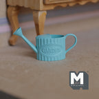 1:12 scale miniature embossed metal watering can in teal with spray nozzle and garden lettering, shown on the floor