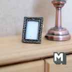 1:12 scale miniature metal rectangular photo frame with stand in antique silver, ornate border, shown beside a lamp