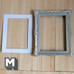 1:12 scale miniature ornate picture frame with mat in silver, large, carved corners, shown with a white mat on a wood surface