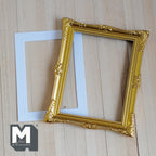 1:12 scale miniature ornate picture frame with mat in gold, large, carved corners, shown with a white mat on a wood surface