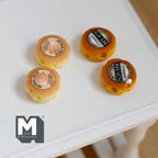 1:12 scale miniature round cheese wheels, set of 4, yellow and orange wheels with printed labels, on a white table