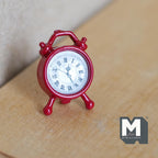 1:12 scale miniature metal twin bell alarm clock in red with a white Roman numeral dial, shown on a wood surface