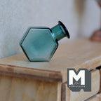1:12 scale miniature glass hexagon bottle vase in teal green, shown lying on its side on a wood dresser
