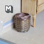 1:12 scale miniature round woven basket with ribbon weave in brown, shown on the floor beside a wood dresser