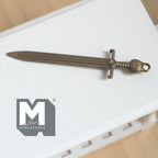 1:12 scale miniature metal sword with ornate hilt in antique bronze, shown lying on a white table