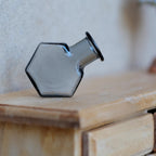 1:12 scale miniature hexagon glass bottle vase in smoky gray, shown lying on its side on a wood dresser
