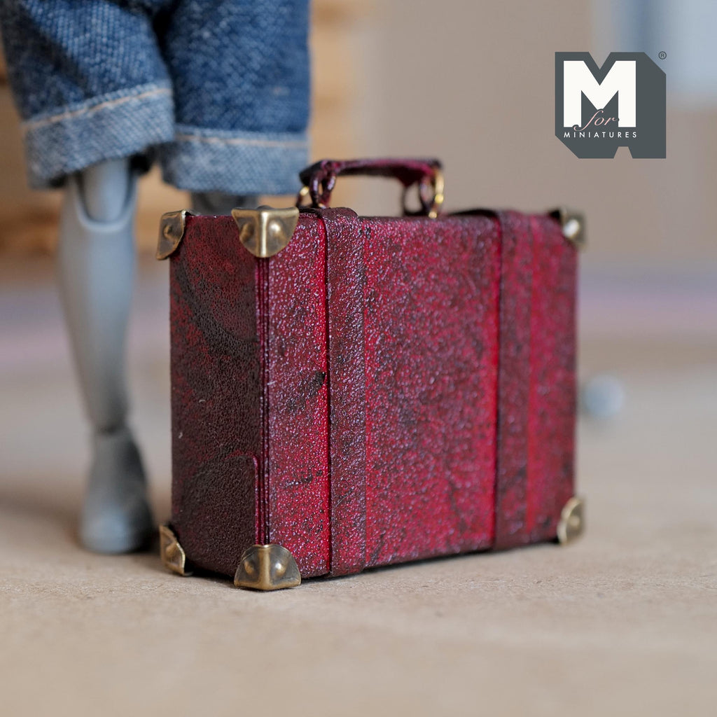 1:12 scale miniature burgundy faux leather suitcase with a worn distressed finish, two buckle straps, metal corner caps and a top handle on metal rings