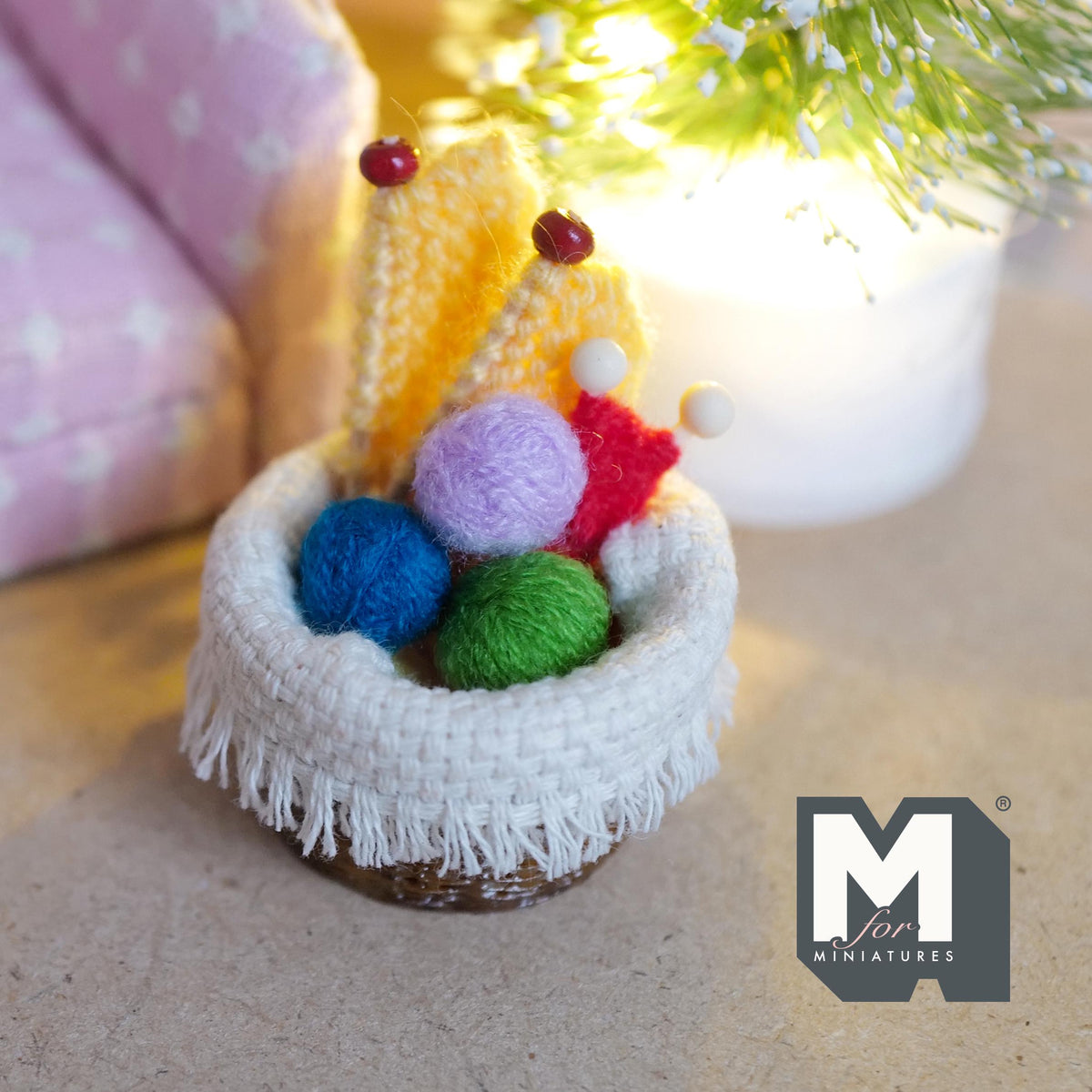 1:12 scale miniature aged woven knitting basket with a fringed cream knit blanket, blue, lilac and green yarn balls and two knitting projects on bead-topped needles