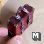 1:12 scale miniature burgundy faux leather suitcase with an aged worn finish, eight antique copper corner caps, two buckle straps and a top handle