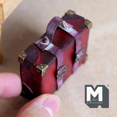 1:12 scale miniature burgundy faux leather suitcase with an aged worn finish, eight antique copper corner caps, two buckle straps and a top handle