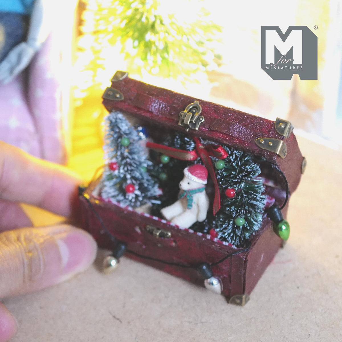 1:12 scale miniature burgundy Christmas storage trunk with brass corners and clasp, lid fixed open, filled with a frosted tree with baubles, wreath with a red bow, bear in a Santa hat and a string of bulb lights