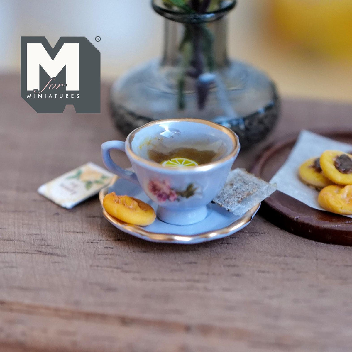 1:12 scale miniature purple cup and saucer with gold rims and a pink rose, filled with lemon tea and a lemon slice, with a tea bag and a jam cookie on the saucer and a lemon tea packet beside it