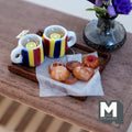 1:12 scale miniature lemon tea tray with two white mugs with red, yellow and blue stripes, lemon slices, a tea bag, and assorted swirl, jam and glazed cookies on baking paper on a wood tray