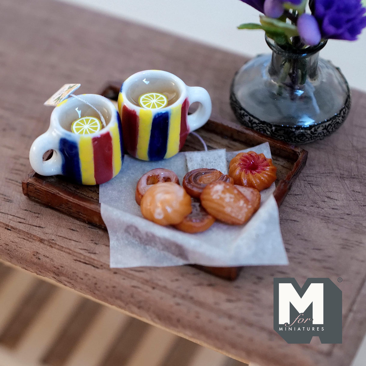 1:12 scale miniature lemon tea tray with two white mugs with red, yellow and blue stripes, lemon slices, a tea bag, and assorted swirl, jam and glazed cookies on baking paper on a wood tray