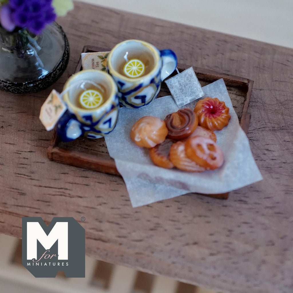 1:12 scale miniature lemon tea tray with two blue and white ceramic mugs with gold rims and lemon slices, a tea bag, and assorted glazed cookies on baking paper on a dark wood tray