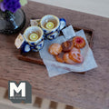 1:12 scale miniature lemon tea tray with two blue and white ceramic mugs with gold rims and lemon slices, a tea bag, and assorted glazed cookies on baking paper on a dark wood tray