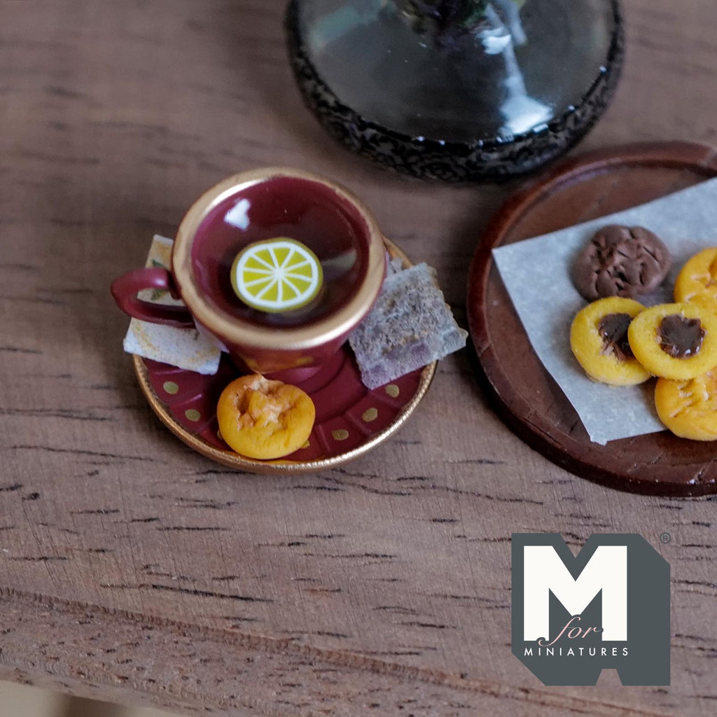 1:12 scale miniature burgundy cup and saucer with gold polka dots and gold rims, filled with tea and a lemon slice, with a tea bag, a tea packet and a jam cookie on the saucer
