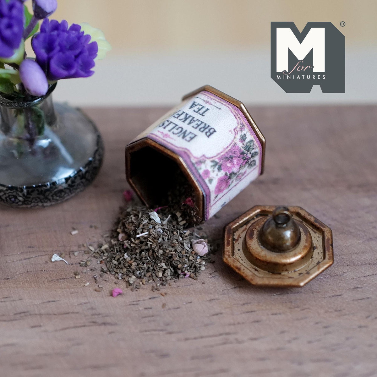 1:12 scale miniature aged metal octagonal tea tin with a pink and purple floral English Breakfast Tea label, lying on its side with loose tea leaves spilling out and its lid with a round cap beside it