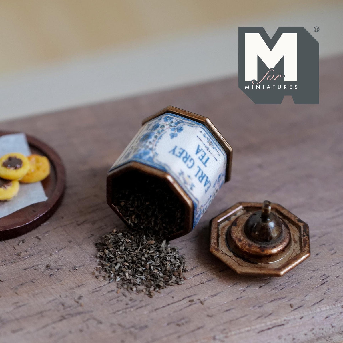 1:12 scale miniature aged metal octagonal tea tin with a blue and white Earl Grey Tea label, lying on its side with loose tea leaves spilling out and its lid with a round cap beside it