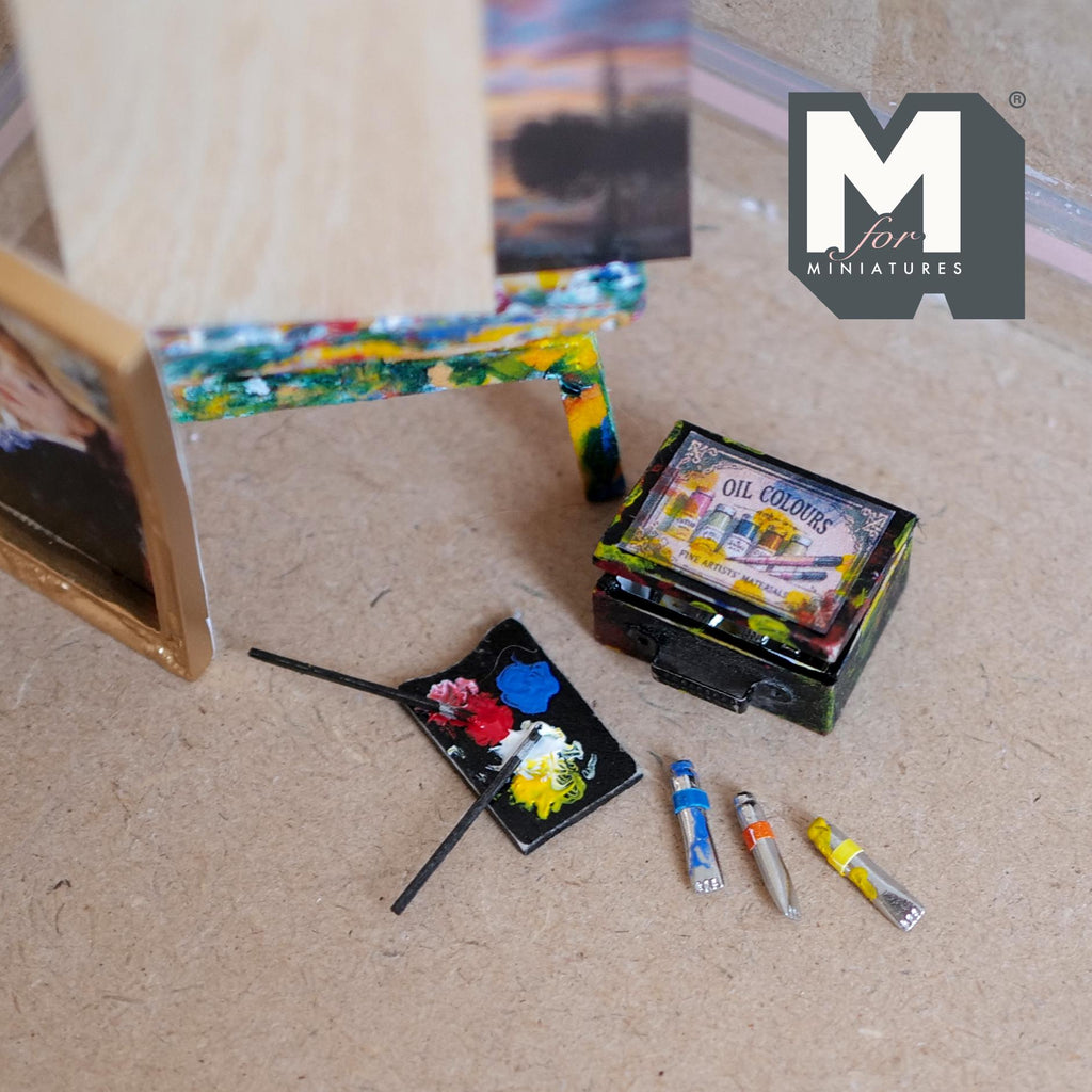 1:12 scale miniature black artist oil paint box splattered with colorful paint and labeled Oil Colors, with loose paint tubes, a brush and a palette with dabs of red, blue, yellow and white paint