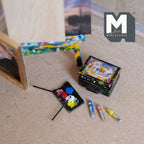 1:12 scale miniature black artist oil paint box splattered with colorful paint and labeled Oil Colors, with loose paint tubes, a brush and a palette with dabs of red, blue, yellow and white paint