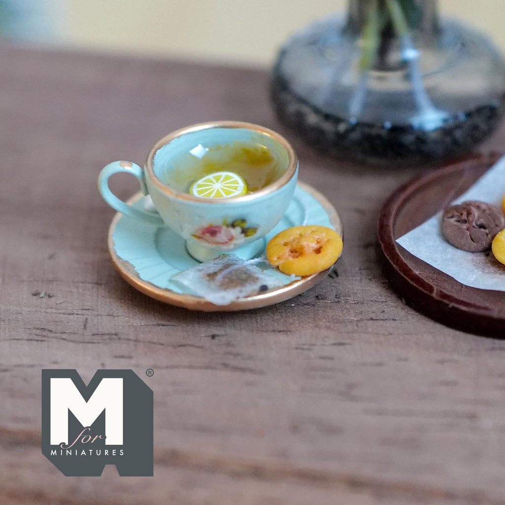 1:12 scale miniature mint green cup and saucer with gold rims and a pink rose, filled with lemon tea and lemon slices, with a tea bag and a jam cookie on the saucer