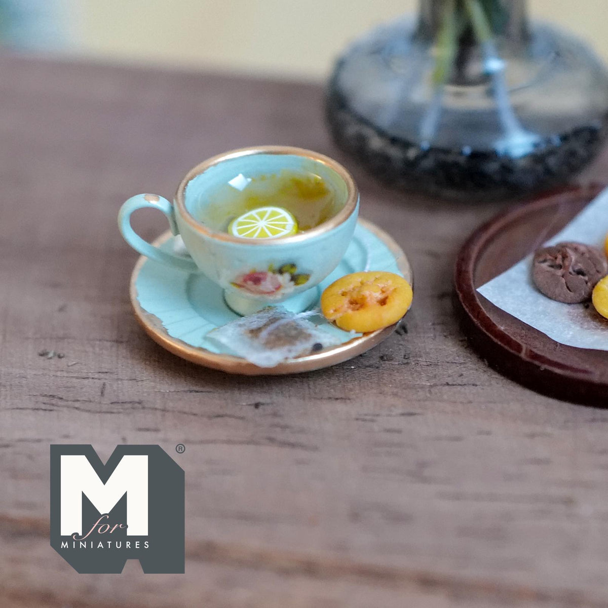 1:12 scale miniature mint green cup and saucer with gold rims and a pink rose, filled with lemon tea and lemon slices, with a tea bag and a jam cookie on the saucer