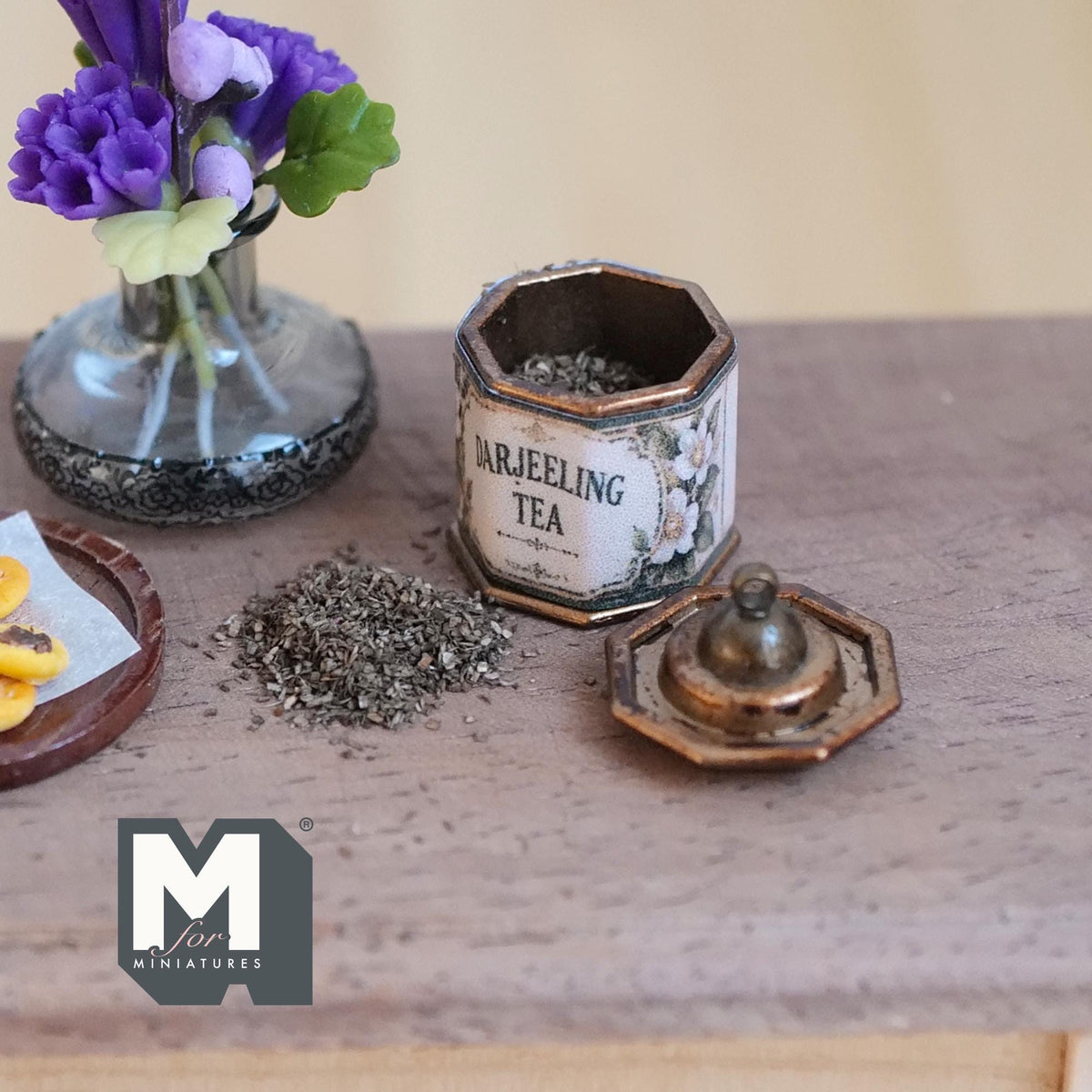 1:12 scale miniature aged metal octagonal tea tin with a white floral Darjeeling Tea label, filled with loose tea leaves, its knobbed lid and a small pile of tea leaves beside it on a wood table