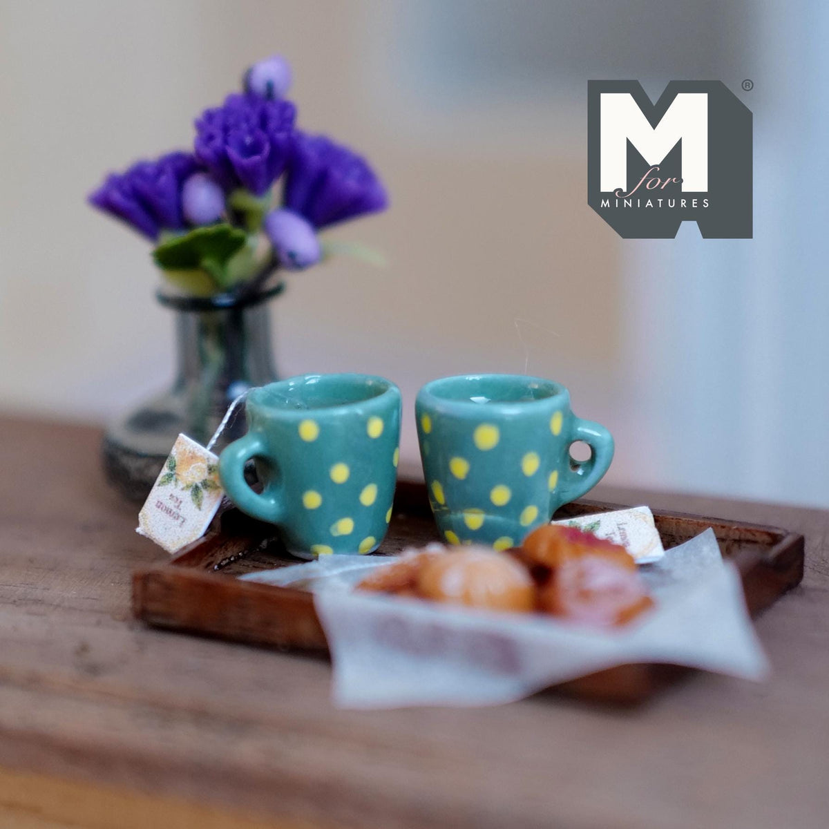 1:12 scale miniature lemon tea tray with two teal mugs with yellow polka dots, a tea tag, and assorted cookies on baking paper on a wood tray, beside a vase of purple flowers
