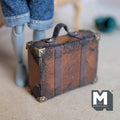1:12 scale miniature brown faux leather suitcase with a worn distressed finish, two wrap-around buckle straps, metal corner caps and a strap handle