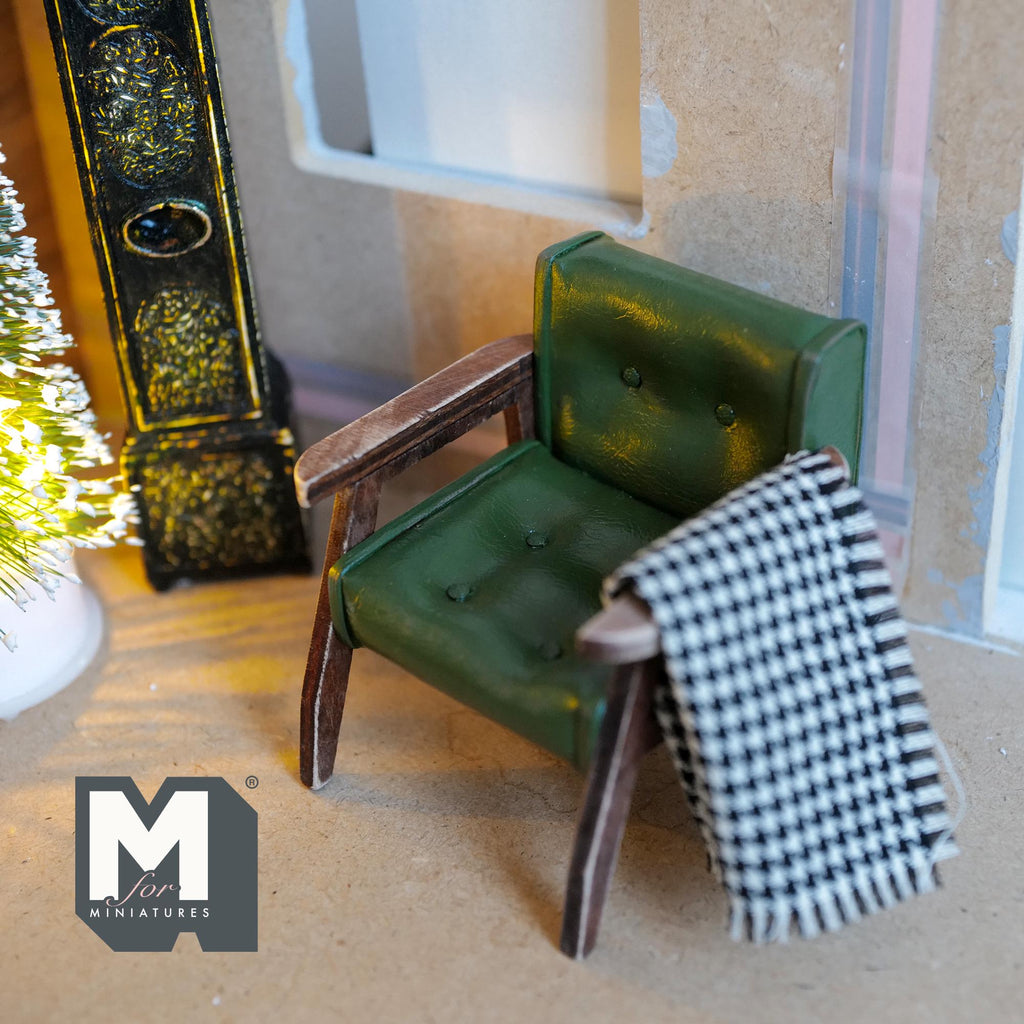1:12 scale miniature armchair with a weathered wood frame, dark green faux leather seat with nail heads, and a black and white checkered blanket over one arm