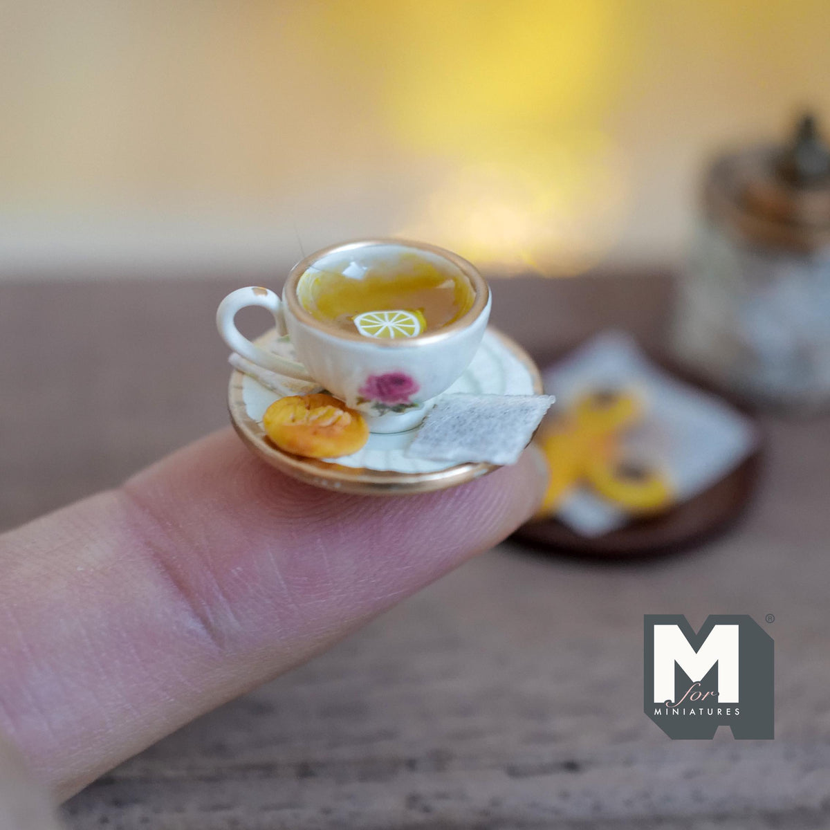 1:12 scale miniature cream tea cup with a pink rose and gold rim, filled with amber tea and lemon slices, on a gold rimmed saucer with a tea bag and jam cookie