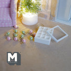 1:12 scale miniature Christmas ornaments in pink and mint green with gold-tone caps, scattered beside a white 12 compartment storage box with a clear lid and a coil of brass wire