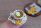 1:12 scale miniature white tea cup with a pink rose and gold rim, filled with lemon tea and a lemon slice, on a gold-rimmed saucer with tea bag and cookie, held on a fingertip