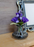 1:12 scale miniature clear glass vase with a dark sparkly base, holding purple carnations, lilac buds and green leaves on a wood shelf