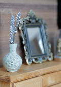 1:12 scale miniature whitewashed round clay vase with raised pebble pattern and silvery white flower stems, next to an ornate framed mirror on a wood dresser