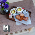 1:12 scale miniature lemon tea tray with two white mugs with red and green polka dots and red handles, lemon slices, a tea bag, and assorted sugar topped and glazed cookies on baking paper