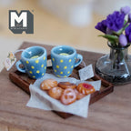 1:12 scale miniature lemon tea tray with two blue mugs with yellow polka dots and lemon slices, a tea tag, and assorted cookies on baking paper on a dark wood tray, beside a vase of purple flowers