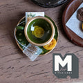 1:12 scale miniature green cup and saucer with gold polka dots and gold rims, seen from above, filled with tea and two lemon slices, with a tea bag and a jam cookie on the saucer