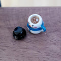 1:12 scale miniature white snowman mug with a blue scarf, filled with hot chocolate and marshmallows, with its black top hat lid beside it on a wood table